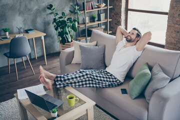 Photo of handsome guy sit sofa legs on table hands behind head work notebook freelancer remote work home quarantine internet job good mood look up pajama pants t-shirt living room indoors