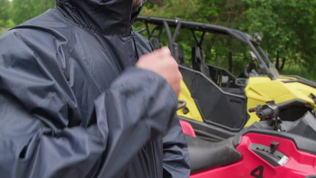 Slowmo tilt up of middle-aged man with beard zipping up his jacket outdoors. Red quad bike and yellow four-wheeler all-terrain vehicle parked beside him