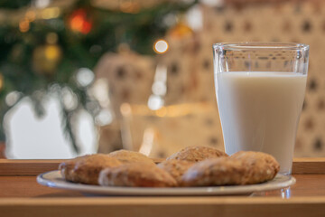 Glass of Milk and Cookies with a christmas tree in a background and also presents in a tree. Gifts for Santa Claus 2020. 