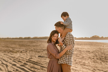 Happy family walking on sandy beach of river. Father, mother holding baby son on hands and going together. Rear view. Family Ties concept.