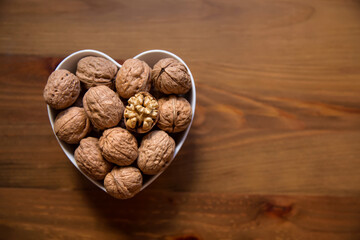 Whole walnuts and one cracked broken heart in a white heart-shaped bowl on a wooden background. Top view. Place for text.