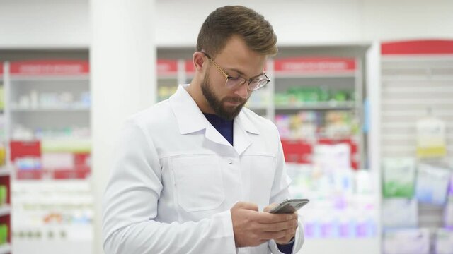 Young caucasian male pharmacist doctor in white medical uniform using smartphene.