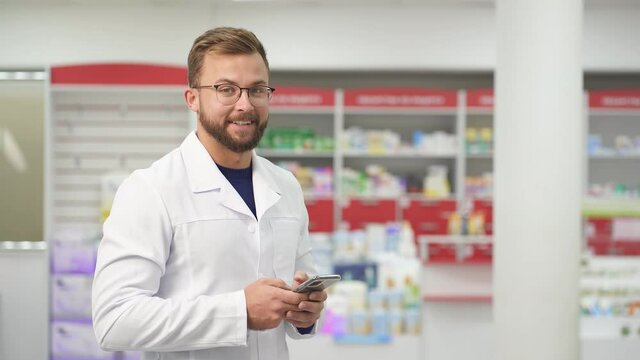 Young caucasian positive male pharmacist doctor in white medical uniform using smartphene, look at camera and smile. portrait