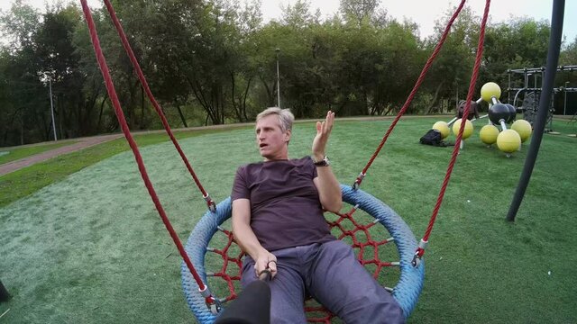 Man swinging on playground and talking at camera