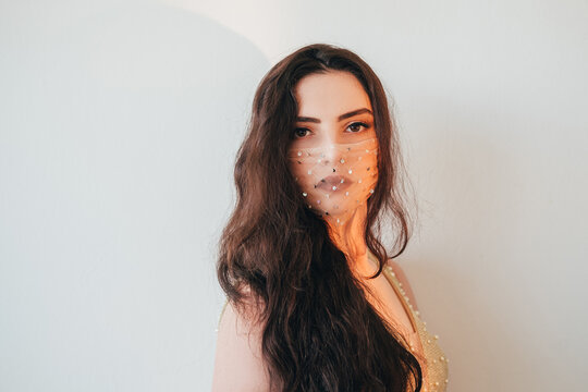 Pandemic beauty. DIY accessory. Fashion trend 2021. Portrait of sensual brunette woman in glamour sequin mesh face mask matching outfit looking at camera on light neutral empty space background.