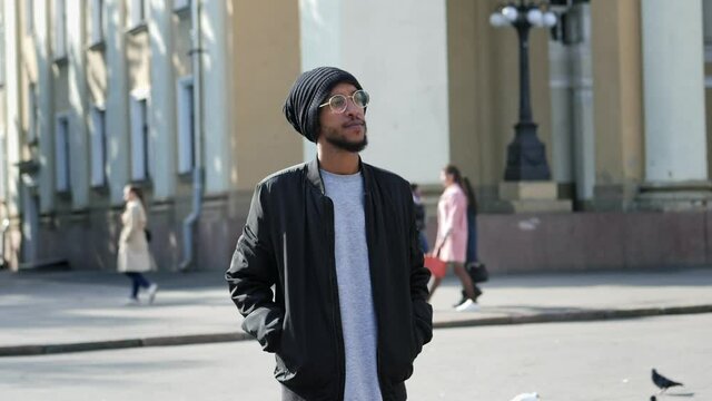 Urban portrait of arab nationality man with glasses, standing on the street dressed traditional hat and looking at the camera in slow motion, people on the background