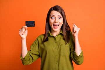 Photo of amazed young lady demonstrate debit card open mouth raise fist wear green shirt isolated orange color background