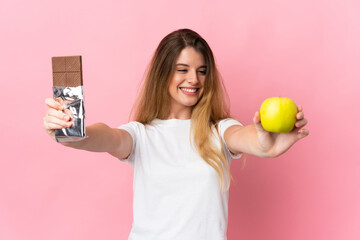 Young blonde woman isolated on pink background taking a chocolate tablet in one hand and an apple in the other