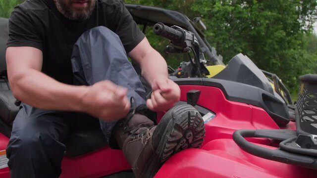 PAN slowmo of unrecognizable bearded man sitting on red quad bike outdoors and tying laces on his boots