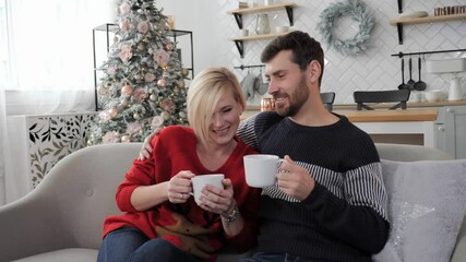 Festive mood, Christmas or New Year, a young couple sitting on the couch and drinking hot coffe. Christmas decor and modern white room. - Powered by Adobe