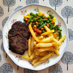 View from above close up of homemade pan seared steak served with french fries and sauteed carrots, corns and peas in a plate on table. Indoor, natural light.