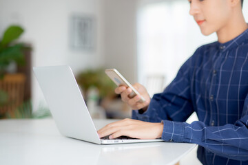 Closeup hand of young man working laptop computer and reading smartphone on internet online on desk at home, freelance male using phone with social media, business and communication concept.