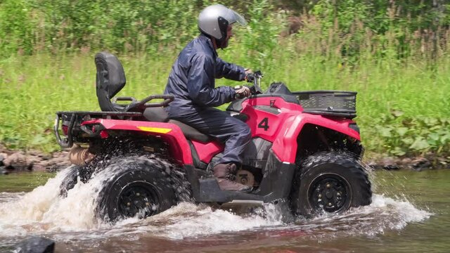 Slow Motion tracking of bearded man in helmet riding red quad bike through river in forest