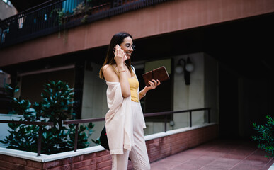 Happy lady with notebook having conversation on smartphone near entrance of building