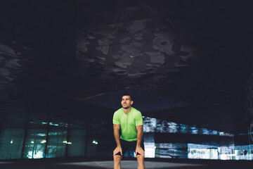 Fototapeta na wymiar Serious male runner standing in dark hall near modern building and resting after training in city