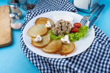 Deruny - potato pancakes with mushrooms, champignons, sour cream on a white plate. Cherry tomatoes, salad. Restaurant serving on a blue background, checkered napkin, fork, knife. Top view, copy space.