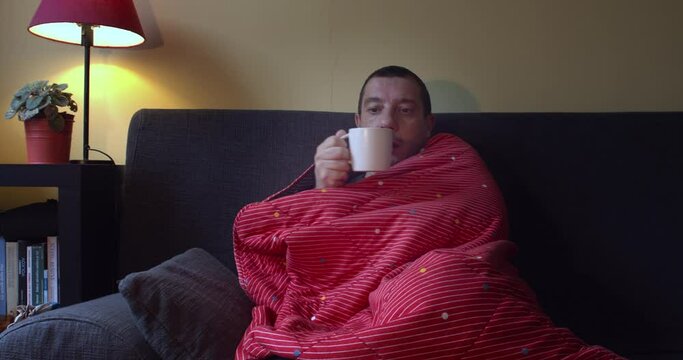 A ill man with flu sitting on the sofa wrapped in a red  blanket while drinking hot tea  