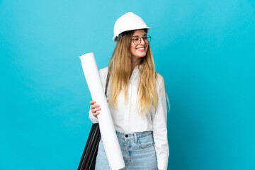 Young architect woman with helmet and holding blueprints isolated on blue background looking side