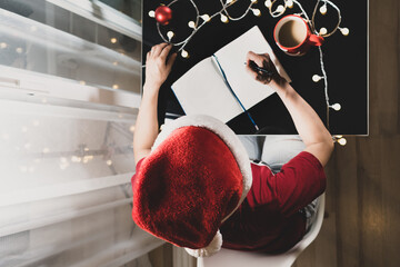 Woman in Santa's hat sitting and writing Christmas shopping list on table while drinking coffee