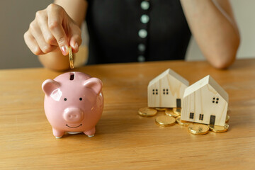 A female housewife putting gold coins into a piggy bank. To save money to buy a new home with model houses as an example of a dream house, family planning ideas, and a new home in the future.