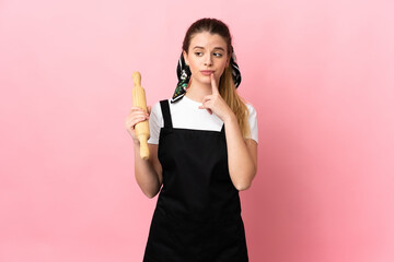 Young blonde woman holding a rolling pin isolated on pink background having doubts while looking up