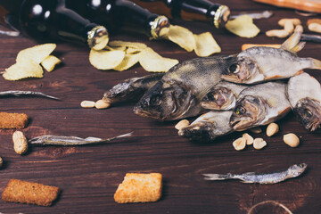 Dried fish, perch, bleak, chips, crackers, nuts and three brown glass bottles of beer on a wooden background.