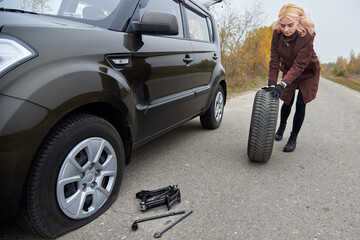 A young blonde woman rolls spare tire near her car with a flat tire.