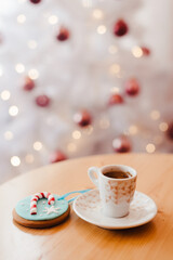 Cup of coffee with ginger bread cookie on the table near the white Christmas tree with golden bokeh lights