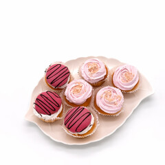 A white porcelain plate in the shape of a leaf filled with pastries. They are decorated with white, pink, raspberry cream, chopped nuts, and chocolate glaze. View from above. Isolated, white backgroun