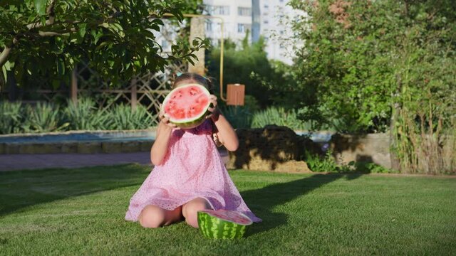 child makes faces, funny small girl fooling around with a watermelon at the camera on a green lawn in the park