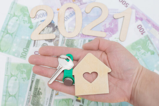 top view, close-up, in hand - a wooden house with a window in the shape of a heart and keys. Against the background of euros and dollars. Concept - buying a house in 2021,planning, ending the mortgage