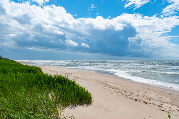Rough sea with waves, cloudy sky, sandy beach and dunes with reeds and green grass