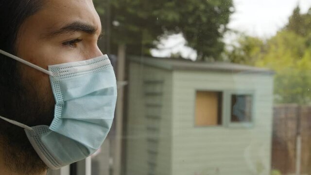 Male looking out of a window longingly putting on a facemask in quarantine close up