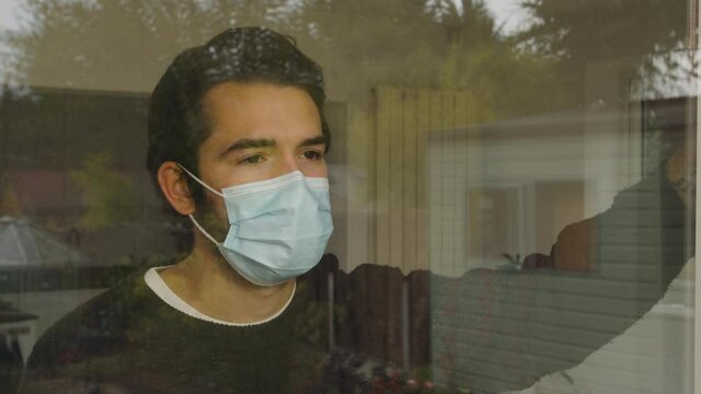 Male looking out and leaning on a window longingly wearing a facemask in quarantine.