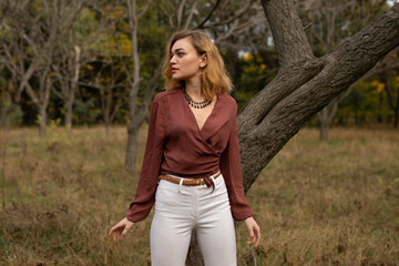 Beautiful blonde woman in in a brown blouse and white trousers posing in the forest