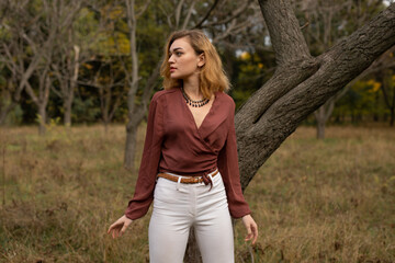 Obraz premium Beautiful blonde woman in in a brown blouse and white trousers posing in the forest