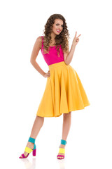 Young Woman In Colorful High Heels, Yellow Skirt And Pink Top Is Showing Peace Hand Sign.