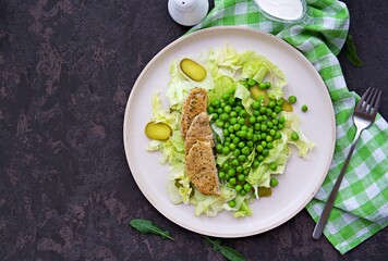 Healthy salad made of iceberg lettuce, fresh green peas, pickled cucumbers and pork on a light ceramic plate on a dark brown concrete background. Salad recipes.