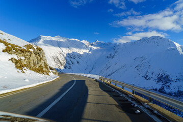 vue depuis une voiture qui roule sur la route d'un col alpin suisse au soleil d'hiver avec des montagnes enneig&eacute;es tout autour.  il n y a pas d autre voiture sur la route