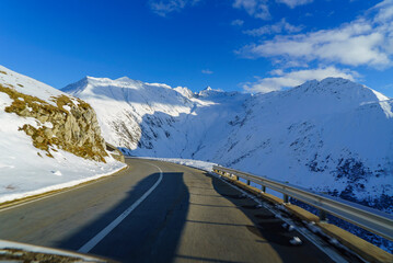 vue depuis une voiture qui roule sur la route d'un col alpin suisse au soleil d'hiver avec des montagnes enneigées tout autour.  il n y a pas d autre voiture sur la route