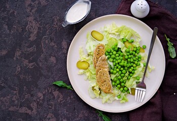 Healthy salad made of iceberg lettuce, fresh green peas, pickled cucumbers and pork on a light ceramic plate on a dark brown concrete background. Salad recipes.
