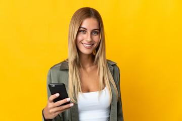 Young blonde woman isolated on yellow background using mobile phone