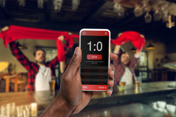 Device screen with mobile app for betting and score. Device with match results on screen, excited fans on background during match. Gambling, betting, sport, finance, modern technologies concept.
