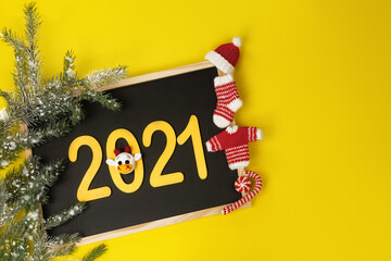 black chalk board with wooden numbers 2021 gold color, fir-tree snow branch and decorative knitted ornaments sock, sweater, hat and caramel in red and white tone on yellow background
