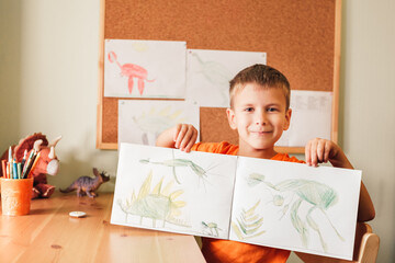 Cute child boy drawing dinosaurs on picture with colored pencils sitting by desk at his room