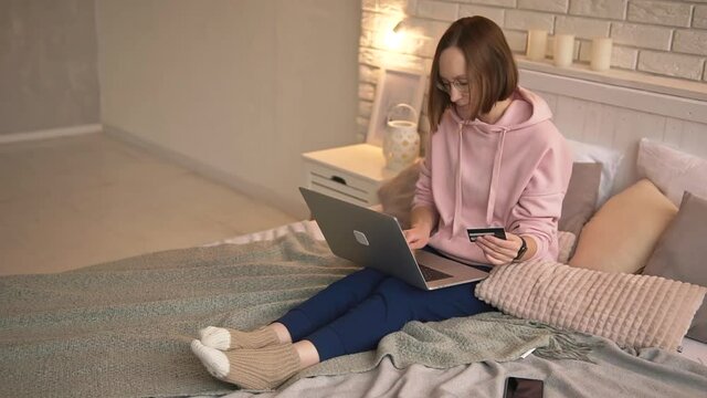 Casual girl shopping online, ordering food, paying bills, transacting with credit card and laptop while sitting at home on her bed. 