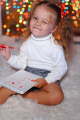 a little girl in a white sweater and shorts with ponytails sits on a white rug against the background of Christmas garlands and holds a Notepad and a marker pen