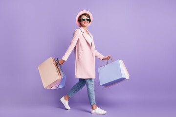 Profile full size photo of cheerful pretty young lady go hold purchases wear glasses coat jeans shoes isolated on purple background
