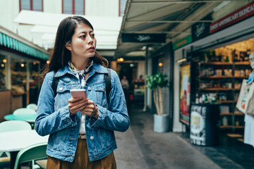 asian woman holding smartphone and looking away is seeking a store. korean lady just moving and living alone overseas is trying to get familiar with the environment. - obrazy, fototapety, plakaty