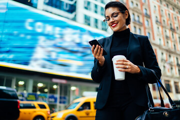 Cheerful young businesswoman standing on street with smartphone and coffee